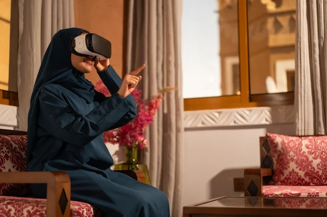 إيماءات التفاعل والاندماج مع الواقع الافتراضي، قضاء أوقات الفراغ بشكل ممتع، امرأة عربية خليجية سعودية ترتدي الحجاب والعباءة تجلس على الاريكة تضع نظارات الفي آر، استخدام الأجهزة التقنية الحديثة والتكنولوجيا في رؤية المشاهد الخيالية