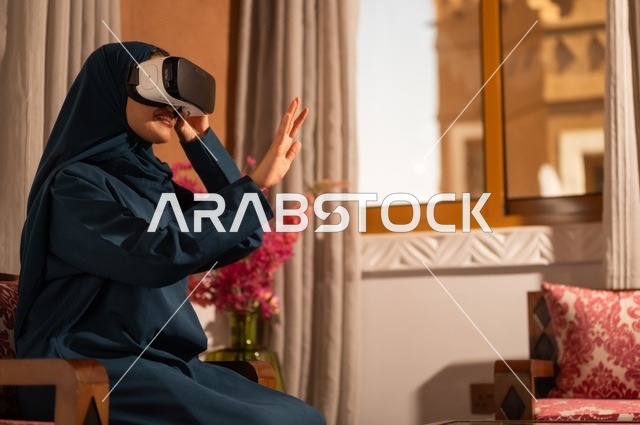 إيماءات التفاعل والاندماج مع الواقع الافتراضي، قضاء أوقات الفراغ بشكل ممتع، امرأة عربية خليجية سعودية ترتدي الحجاب والعباءة تجلس على الاريكة تضع نظارات الفي آر، استخدام الأجهزة التقنية الحديثة والتكنولوجيا في رؤية المشاهد الخيالية