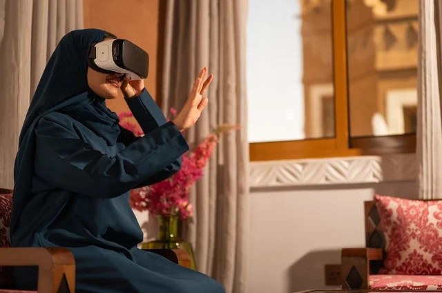 إيماءات التفاعل والاندماج مع الواقع الافتراضي، قضاء أوقات الفراغ بشكل ممتع، امرأة عربية خليجية سعودية ترتدي الحجاب والعباءة تجلس على الاريكة تضع نظارات الفي آر، استخدام الأجهزة التقنية الحديثة والتكنولوجيا في رؤية المشاهد الخيالية