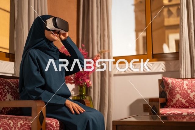 Gestures of interaction and immersion with virtual reality, spending leisure time in an enjoyable way, an Arab Gulf Saudi woman wearing a hijab and abaya sitting on the sofa wearing VR glasses, using modern technological devices and technology to see imaginary scenes.