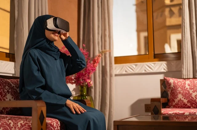 إيماءات التفاعل والاندماج مع الواقع الافتراضي، قضاء أوقات الفراغ بشكل ممتع، امرأة عربية خليجية سعودية ترتدي الحجاب والعباءة تجلس على الاريكة تضع نظارات الفي آر، استخدام الأجهزة التقنية الحديثة والتكنولوجيا في رؤية المشاهد الخيالية