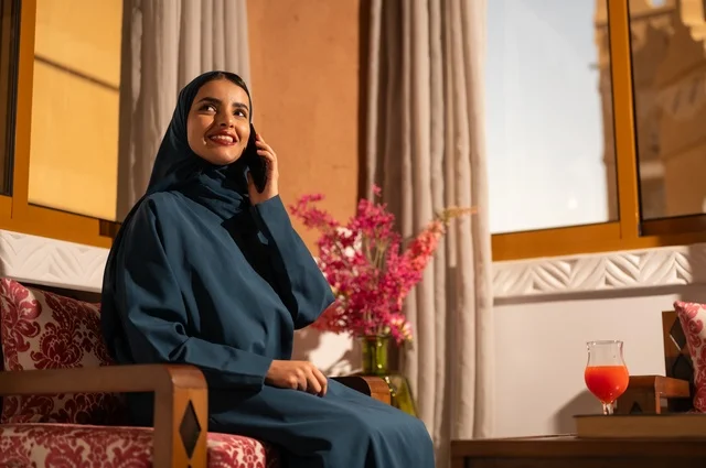 قضاء اوقات ممتعة بتبادل الاخبار والأحاديث، التحدث عبر الهاتف المحمول، بورتريه لامرأة عربية خليجية سعودية محجبة ترتدي العباءة تجلس على الاريكة في غرفة الجلوس تقوم بإجرء مكالمة هاتفية، محادثات ودردشات مع الاصدقاء