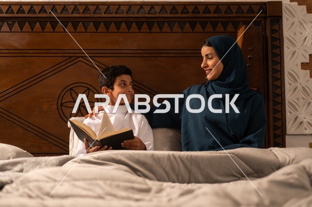 قراءه القصص المفيده للأبناء قبل النوم، مفهوم العطف والحنان عند الأمهات، تربية الأبناء والاهتمام بهم، تقوية روابط المحبة والتماسك الأسري، أم عربية خليجية سعودية محجبة تجلس على سرير مريح برفقة ولدها تقوم بقراءة قصة مسلية، ايماءات السعادة والسرور