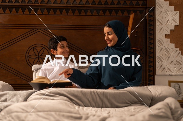 قراءه القصص المفيده للأبناء قبل النوم، مفهوم العطف والحنان عند الأمهات، تربية الأبناء والاهتمام بهم، تقوية روابط المحبة والتماسك الأسري، أم عربية خليجية سعودية محجبة تجلس على سرير مريح برفقة ولدها تقوم بقراءة قصة مسلية، ايماءات السعادة والسرور