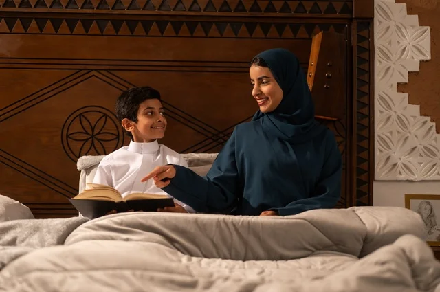 قراءه القصص المفيده للأبناء قبل النوم، مفهوم العطف والحنان عند الأمهات، تربية الأبناء والاهتمام بهم، تقوية روابط المحبة والتماسك الأسري، أم عربية خليجية سعودية محجبة تجلس على سرير مريح برفقة ولدها تقوم بقراءة قصة مسلية، ايماءات السعادة والسرور