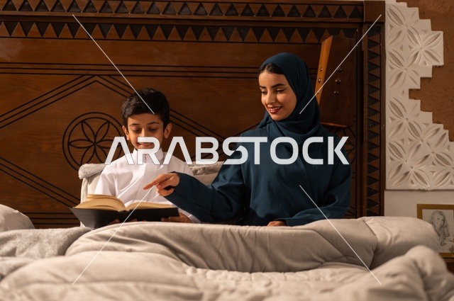 قراءه القصص المفيده للأبناء قبل النوم، مفهوم العطف والحنان عند الأمهات، تربية الأبناء والاهتمام بهم، تقوية روابط المحبة والتماسك الأسري، أم عربية خليجية سعودية محجبة تجلس على سرير مريح برفقة ولدها تقوم بقراءة قصة مسلية، ايماءات السعادة والسرور