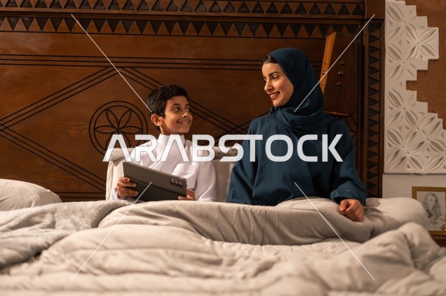 Reading beneficial stories to children before bedtime, the concept of affection and tenderness among mothers, raising children and caring for them, strengthening the bonds of love and family cohesion, an Arab Gulf Saudi mother in a hijab sitting on a comfortable bed with her son reading an entertaining story, gestures of happiness and joy.