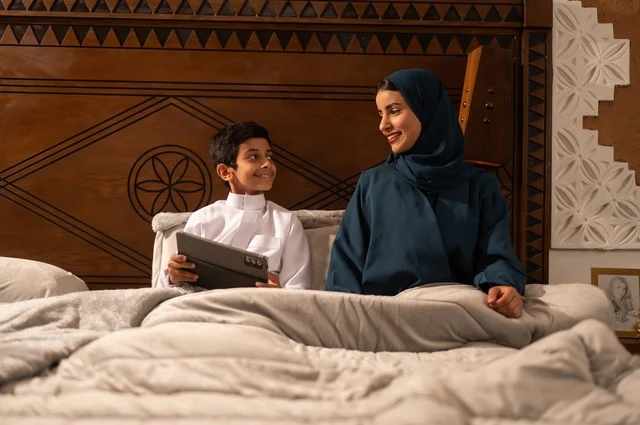 Reading beneficial stories to children before bedtime, the concept of affection and tenderness among mothers, raising children and caring for them, strengthening the bonds of love and family cohesion, an Arab Gulf Saudi mother in a hijab sitting on a comfortable bed with her son reading an entertaining story, gestures of happiness and joy.