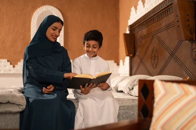 قراءه القصص المفيده للأبناء قبل النوم، مفهوم العطف والحنان عند الأمهات، تربية الأبناء والاهتمام بهم، تقوية روابط المحبة والتماسك الأسري، أم عربية خليجية سعودية محجبة تجلس على سرير مريح برفقة ولدها تقوم بقراءة قصة مسلية، ايماءات السعادة والسرور