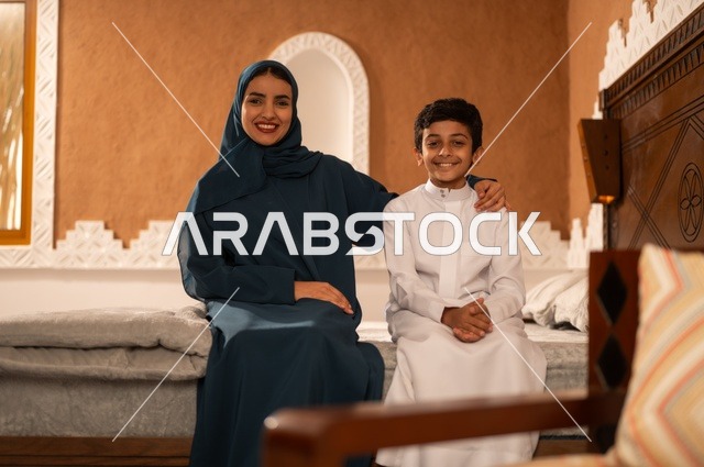 مفهوم العطف والحنان عند الأمهات، تربية الأبناء والاهتمام بهم، تقوية روابط المحبة والتماسك الأسري، الاستمتاع بقضاء الوقت مع الأبناء،أم عربية خليجية سعودية محجبة تجلس مع ابنها في غرفة الجلوس، النظر إلى الكاميرا بايماءات السعادة والسرور