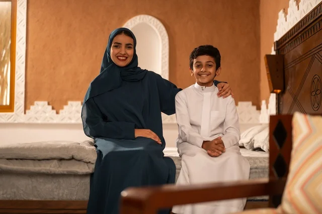 مفهوم العطف والحنان عند الأمهات، تربية الأبناء والاهتمام بهم، تقوية روابط المحبة والتماسك الأسري، الاستمتاع بقضاء الوقت مع الأبناء،أم عربية خليجية سعودية محجبة تجلس مع ابنها في غرفة الجلوس، النظر إلى الكاميرا بايماءات السعادة والسرور