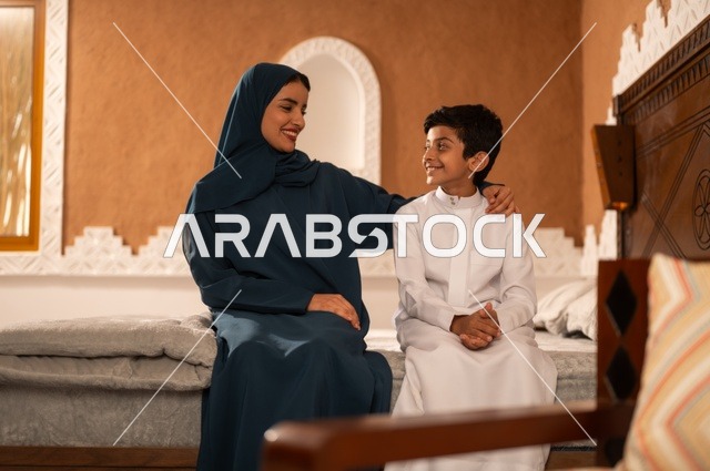 تربية الأبناء والاهتمام بهم، تقوية روابط المحبة والتماسك الأسري، الاستمتاع بقضاء الوقت مع الابناء،أم عربية خليجية سعودية محجبة تجلس مع ابنها في غرفة الجلووس، مفهوم العطف والحنان عند الأمهات، ايماءات السعادة والسرور