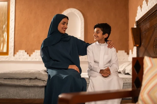 Raising children and caring for them, strengthening the bonds of love and family cohesion, enjoying spending time with children, a Gulf Arab Saudi mother in a hijab sitting with her son in the living room, the concept of affection and tenderness among mothers, gestures of happiness and joy.