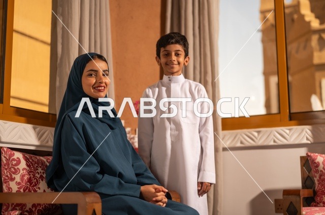 مفهوم العطف والحنان عند الأمهات، تربية الأبناء والاهتمام بهم، تقوية روابط المحبة والتماسك الأسري، الاستمتاع بقضاء الوقت مع الأبناء،أم عربية خليجية سعودية محجبة تجلس مع ابنها في غرفة الجلوس، النظر إلى الكاميرا بايماءات السعادة والسرور