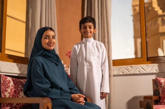 The concept of affection and tenderness among mothers, raising children and caring for them, strengthening the bonds of love and family cohesion, enjoying spending time with children, an Arab Gulf Saudi mother in a hijab sitting with her son in the living room, looking at the camera with gestures of happiness and joy.