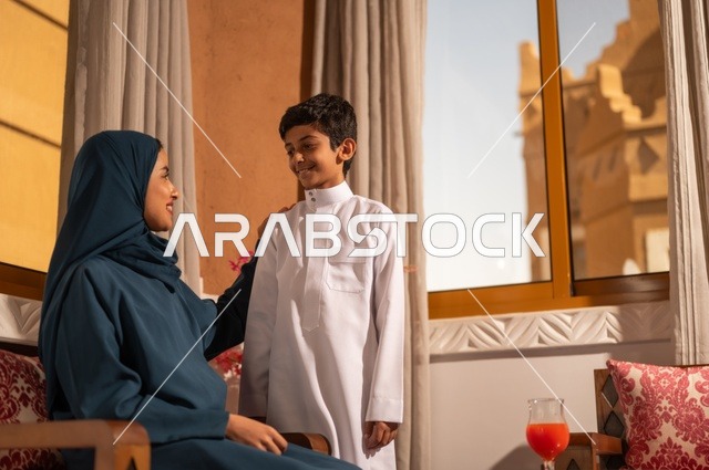 تربية الأبناء والاهتمام بهم، تقوية روابط المحبة والتماسك الأسري، الاستمتاع بقضاء الوقت مع الابناء،أم عربية خليجية سعودية محجبة تجلس مع ابنها في غرفة الجلووس، مفهوم العطف والحنان عند الأمهات، ايماءات السعادة والسرور