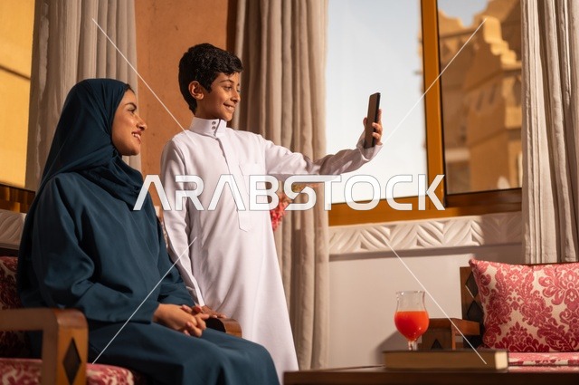 التقاط صورة سيلفي تذكارية باستخدام الهاتف المحمول، مفهوم العطف والحنان عند الأمهات، تربية الأبناء والاهتمام بهم، تقوية روابط المحبة والتماسك الأسري، الاستمتاع بقضاء الوقت مع الابناء،أم عربية خليجية سعودية محجبة تجلس مع ابنها، ايماءات السعادة والسرور