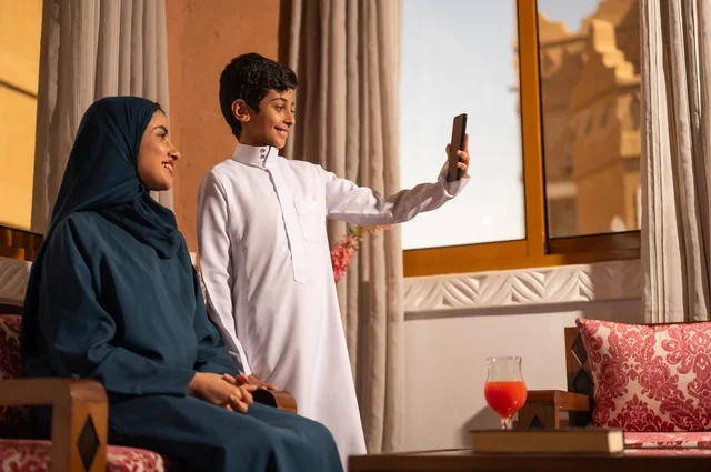 التقاط صورة سيلفي تذكارية باستخدام الهاتف المحمول، مفهوم العطف والحنان عند الأمهات، تربية الأبناء والاهتمام بهم، تقوية روابط المحبة والتماسك الأسري، الاستمتاع بقضاء الوقت مع الابناء،أم عربية خليجية سعودية محجبة تجلس مع ابنها، ايماءات السعادة والسرور