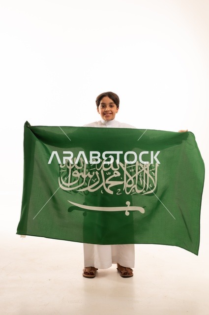 Looking at the camera with gestures of happiness and joy, the memory of Saudi National Day on September 23, pride and honor in possessing national identity, love for the homeland and belonging to it, a portrait of an Arab Gulf Saudi boy wearing traditional attire holding the Kingdom's flag, celebrating Flag Day on March 11, with a white background.