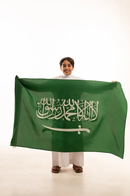 Looking at the camera with gestures of happiness and joy, the memory of Saudi National Day on September 23, pride and honor in possessing national identity, love for the homeland and belonging to it, a portrait of an Arab Gulf Saudi boy wearing traditional attire holding the Kingdom's flag, celebrating Flag Day on March 11, with a white background.