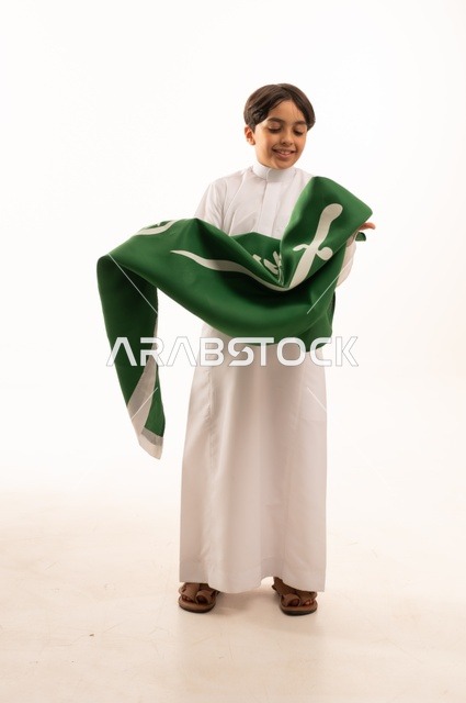 Gestures of happiness and joy, the anniversary of the Saudi National Day on September 23, pride and honor in possessing national identity, love for the homeland and belonging to it, a portrait of an Arab Gulf Saudi boy wearing traditional attire holding the flag of the Kingdom in his hand, celebrating Flag Day on March 11, white background.