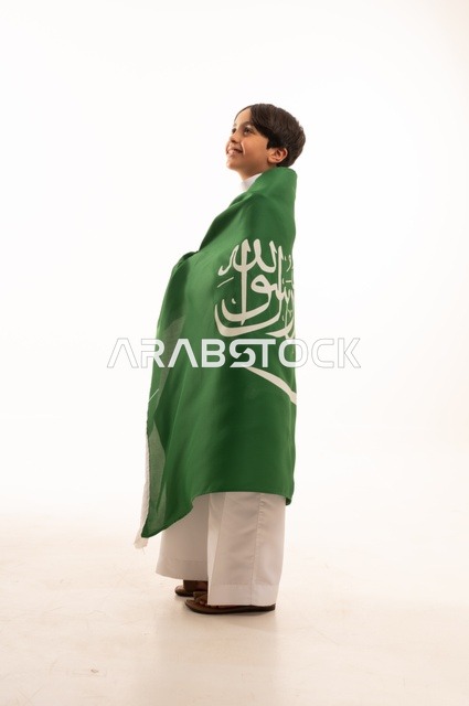 Gestures of happiness and joy, the anniversary of the Saudi National Day on September 23, pride and honor in possessing national identity, love for the homeland and belonging to it, a portrait of an Arab Gulf Saudi boy wearing traditional attire holding the flag of the Kingdom in his hand, celebrating Flag Day on March 11, white background.