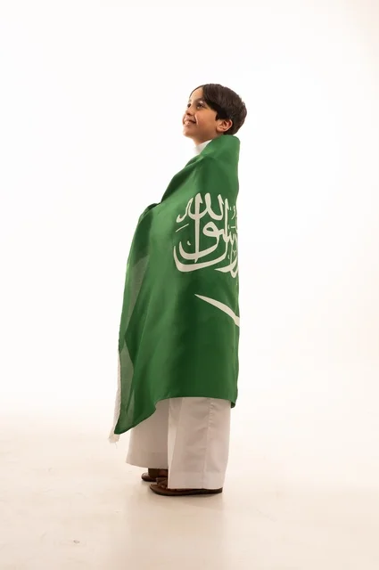 Gestures of happiness and joy, the anniversary of the Saudi National Day on September 23, pride and honor in possessing national identity, love for the homeland and belonging to it, a portrait of an Arab Gulf Saudi boy wearing traditional attire holding the flag of the Kingdom in his hand, celebrating Flag Day on March 11, white background.