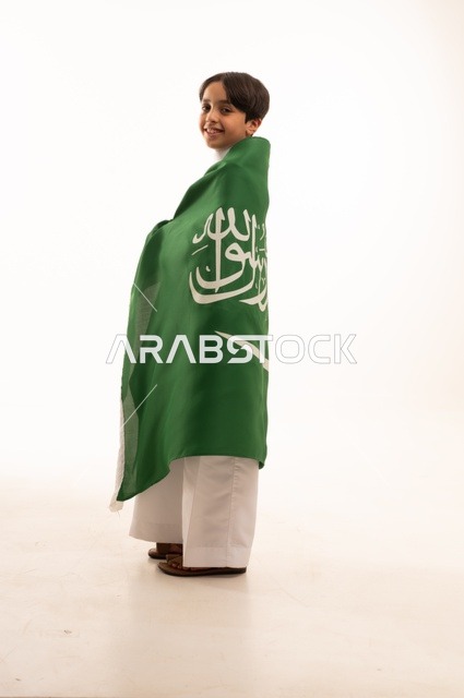 Looking at the camera with gestures of happiness and joy, the memory of Saudi National Day on September 23, pride and honor in possessing national identity, love for the homeland and belonging to it, a portrait of an Arab Gulf Saudi boy wearing traditional attire holding the Kingdom's flag, celebrating Flag Day on March 11, with a white background.