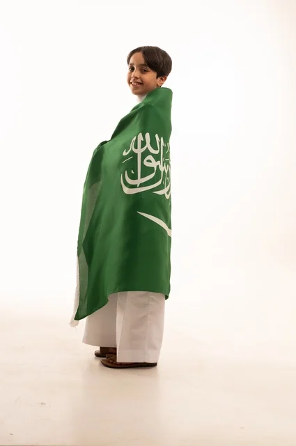 Looking at the camera with gestures of happiness and joy, the memory of Saudi National Day on September 23, pride and honor in possessing national identity, love for the homeland and belonging to it, a portrait of an Arab Gulf Saudi boy wearing traditional attire holding the Kingdom's flag, celebrating Flag Day on March 11, with a white background.