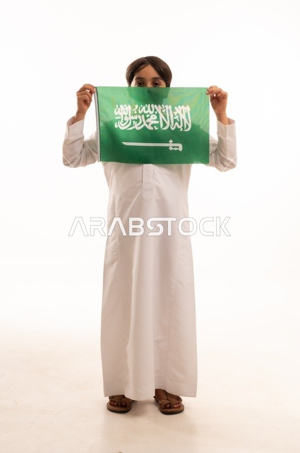 النظر إلى الكاميرا بإيماءات السعادة والسرور، ذكرى اليوم الوطني السعودي 23سبتمبر، الفخر والاعتزاز بامتلاك الهوية الوطنية، حب الوطن والانتماء إليه، بورتريه لولد عربي خليجي سعودي يرتدي الثوب التقليدي يحمل بيده علم المملكة  الاحتفال بيوم العلم 11مارس، خلفية بيضاء
