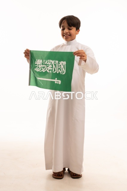 ذكرى اليوم الوطني السعودي 23سبتمبر، الفخر والاعتزاز بامتلاك الهوية الوطنية، حب الوطن والانتماء إليه، بورتريه لولد عربي خليجي سعودي يرتدي الثوب التقليدي يحمل بيده علم المملكة وينظر إلى شيء ما، الاحتفال بيوم العلم 11مارس، خلفية بيضاء