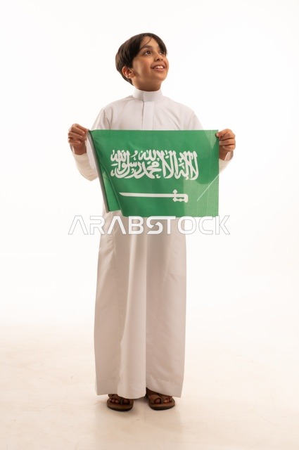 ذكرى اليوم الوطني السعودي 23سبتمبر، الفخر والاعتزاز بامتلاك الهوية الوطنية، حب الوطن والانتماء إليه، بورتريه لولد عربي خليجي سعودي يرتدي الثوب التقليدي يحمل بيده علم المملكة وينظر إلى شيء ما، الاحتفال بيوم العلم 11مارس، خلفية بيضاء