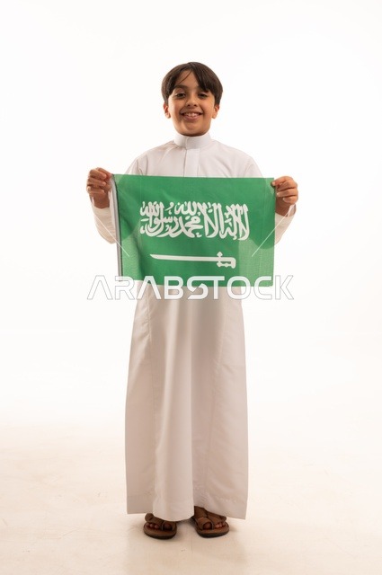 النظر إلى الكاميرا بإيماءات السعادة والسرور، ذكرى اليوم الوطني السعودي 23سبتمبر، الفخر والاعتزاز بامتلاك الهوية الوطنية، حب الوطن والانتماء إليه، بورتريه لولد عربي خليجي سعودي يرتدي الثوب التقليدي يحمل بيده علم المملكة  الاحتفال بيوم العلم 11مارس، خلفية بيضاء