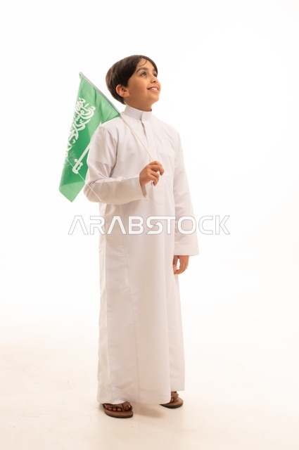 The Saudi National Day is on September 23, a pride and honor in possessing national identity, love for the homeland and belonging to it. A portrait of a young Arab Gulf Saudi boy wearing traditional attire, holding the Kingdom's flag in his hand and looking at something. The celebration of Flag Day is on March 11, with a white background.