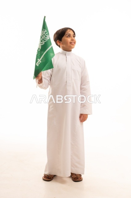 The Saudi National Day is on September 23, a pride and honor in possessing national identity, love for the homeland and belonging to it. A portrait of a young Arab Gulf Saudi boy wearing traditional attire, holding the Kingdom's flag in his hand and looking at something. The celebration of Flag Day is on March 11, with a white background.