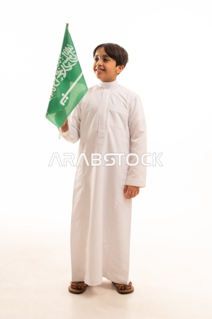 The Saudi National Day is on September 23, a pride and honor in possessing national identity, love for the homeland and belonging to it. A portrait of a young Arab Gulf Saudi boy wearing traditional attire, holding the Kingdom's flag in his hand and looking at something. The celebration of Flag Day is on March 11, with a white background.