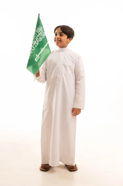The Saudi National Day is on September 23, a pride and honor in possessing national identity, love for the homeland and belonging to it. A portrait of a young Arab Gulf Saudi boy wearing traditional attire, holding the Kingdom's flag in his hand and looking at something. The celebration of Flag Day is on March 11, with a white background.