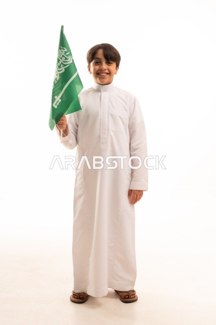 Looking at the camera with gestures of happiness and joy, the memory of Saudi National Day on September 23, pride and honor in possessing national identity, love for the homeland and belonging to it, a portrait of an Arab Gulf Saudi boy wearing traditional attire holding the Kingdom's flag, celebrating Flag Day on March 11, with a white background.