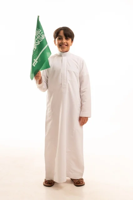 النظر إلى الكاميرا بإيماءات السعادة والسرور، ذكرى اليوم الوطني السعودي 23سبتمبر، الفخر والاعتزاز بامتلاك الهوية الوطنية، حب الوطن والانتماء إليه، بورتريه لولد عربي خليجي سعودي يرتدي الثوب التقليدي يحمل بيده علم المملكة  الاحتفال بيوم العلم 11مارس، خلفية بيضاء