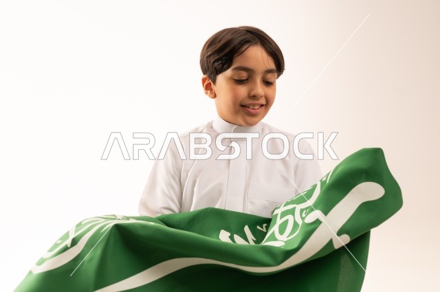 إيماءات السعادة والسرور، ذكرى اليوم الوطني السعودي 23سبتمبر، الفخر والاعتزاز بامتلاك الهوية الوطنية، حب الوطن والانتماء إليه، بورتريه مقرب لولد عربي خليجي سعودي يرتدي الثوب التقليدي يحمل بيده علم المملكة ، الاحتفال بيوم العلم 11مارس، خلفية بيضاء