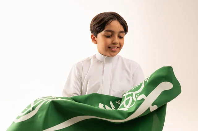 إيماءات السعادة والسرور، ذكرى اليوم الوطني السعودي 23سبتمبر، الفخر والاعتزاز بامتلاك الهوية الوطنية، حب الوطن والانتماء إليه، بورتريه مقرب لولد عربي خليجي سعودي يرتدي الثوب التقليدي يحمل بيده علم المملكة ، الاحتفال بيوم العلم 11مارس، خلفية بيضاء