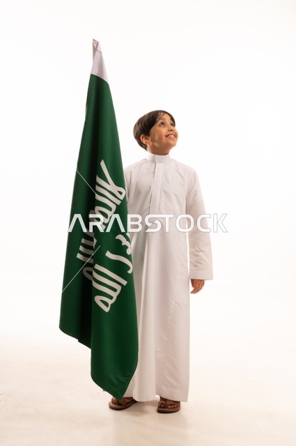 Celebrating the Day of Bidayna 1727, the anniversary of the Saudi National Day on September 23, looking up with gestures of pride and honor in belonging to the homeland, a portrait of an Arab Gulf Saudi boy wearing traditional attire holding in his hand the flag of the establishment of the Saudi Kingdom on February 22, full-body, white background.