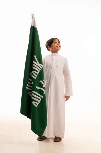 الاحتفال بيوم بدينا1727 ، ذكرى اليوم الوطني السعودي 23سبتمبر، النظر إلى الأعلى بإيماءات الفخر والاعتزاز باالانتماء للوطن، بورتريه لولد عربي خليجي سعودي يرتدي الثوب التقليدي يحمل بيده علم تأسيس المملكة السعودية 22 فبراير، الجسم بالطول الكامل، خلفية بيضاء