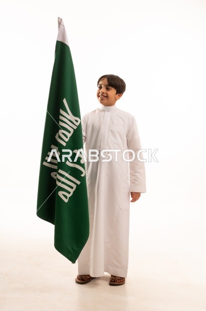 Celebrating the Day of Bidayna 1727, the anniversary of the Saudi National Day on September 23, looking up with gestures of pride and honor in belonging to the homeland, a portrait of an Arab Gulf Saudi boy wearing traditional attire holding in his hand the flag of the establishment of the Saudi Kingdom on February 22, full-body, white background.