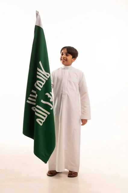 الاحتفال بيوم بدينا1727 ، ذكرى اليوم الوطني السعودي 23سبتمبر، النظر إلى الأعلى بإيماءات الفخر والاعتزاز باالانتماء للوطن، بورتريه لولد عربي خليجي سعودي يرتدي الثوب التقليدي يحمل بيده علم تأسيس المملكة السعودية 22 فبراير، الجسم بالطول الكامل، خلفية بيضاء