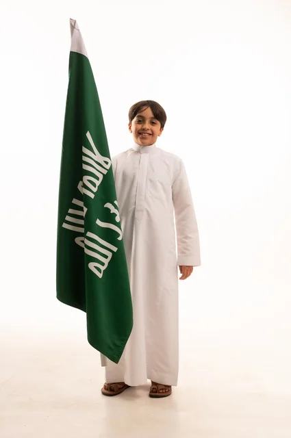 Saudi National Day is celebrated on September 23, looking at the camera with gestures of pride and belonging to the homeland, a portrait of an Arab Gulf Saudi boy wearing traditional attire holding the flag of the establishment of the Saudi Kingdom on February 22, celebrating the day we began in 1727, full-body shot, white background.