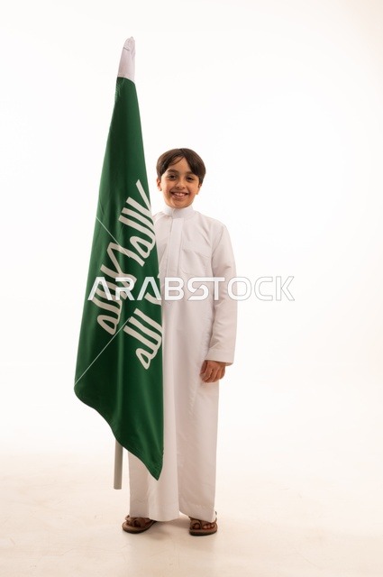 ذكرى اليوم الوطني السعودي 23سبتمبر، النظر إلى الكاميرا بإيماءات الفخر والاعتزاز باالانتماء للوطن، بورتريه لولد عربي خليجي سعودي يرتدي الثوب التقليدي يحمل بيده علم تأسيس المملكة السعودية 22 فبراير، الاحتفال بيوم بدينا1727 ، الجسم بالطول الكامل، خلفية بيضاء