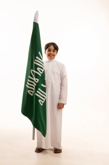 Saudi National Day is celebrated on September 23, looking at the camera with gestures of pride and belonging to the homeland, a portrait of an Arab Gulf Saudi boy wearing traditional attire holding the flag of the establishment of the Saudi Kingdom on February 22, celebrating the day we began in 1727, full-body shot, white background.