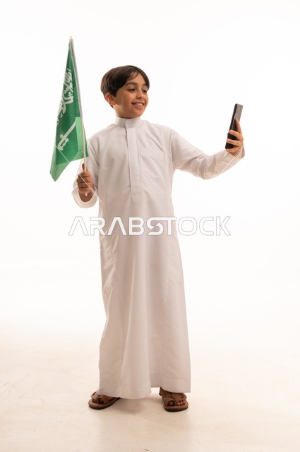 Taking a commemorative selfie using the phone, marking the Saudi National Day on September 23, pride and honor in belonging to the homeland, a portrait of an Arab Gulf Saudi boy wearing traditional attire holding the Kingdom's flag in his hand, celebrating Flag Day on March 11, full-body shot, white background.