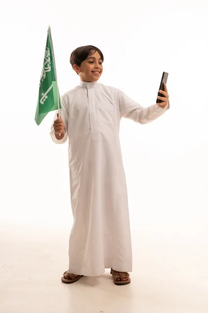 التقاط صورة سيلفي تذكارية باستخدام الهاتف، ذكرى اليوم الوطني السعودي 23سبتمبر، الفخر والاعتزاز باالانتماء للوطن، بورتريه لولد عربي خليجي سعودي يرتدي الثوب التقليدي يحمل بيده علم المملكة ، الاحتفال بيوم العلم 11مارس، الجسم بالطول الكامل، خلفية بيضاء